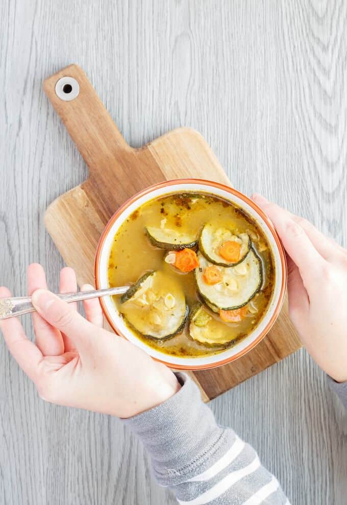 Roasted Zucchini-Carrot Soup in white orange bowl with spoon held by hand on wooden pad