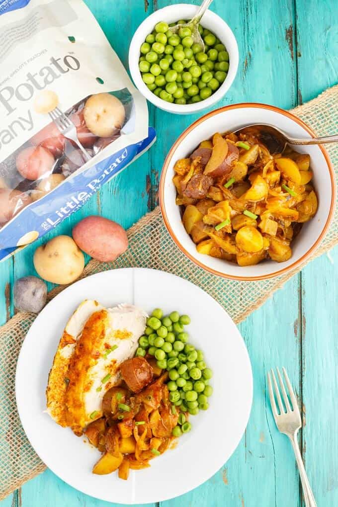 Slow cooker curry chicked on white palte with potatoes and peas on blue table with fork, bowl of potatoes with spoon, bowls of peas and sack of spilled potatoes