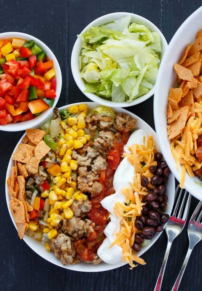 Ground Turkey Taco Salad on white plate with bowls full of vegetable, fruits and chips with forks