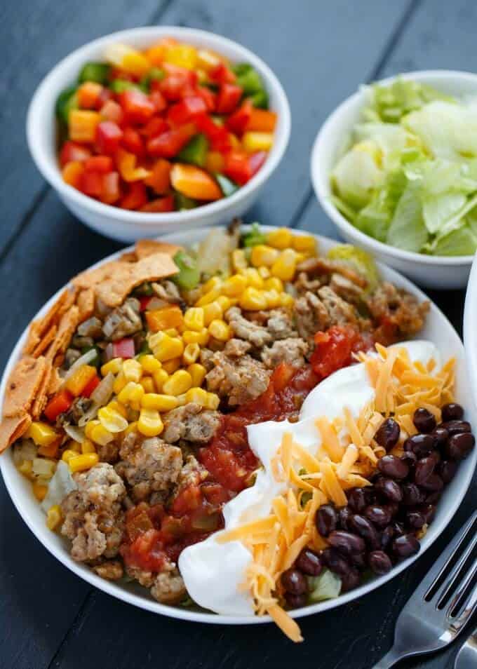 Ground turkey taco salad on white plate with bowls of salad and vegetables on black table with forks