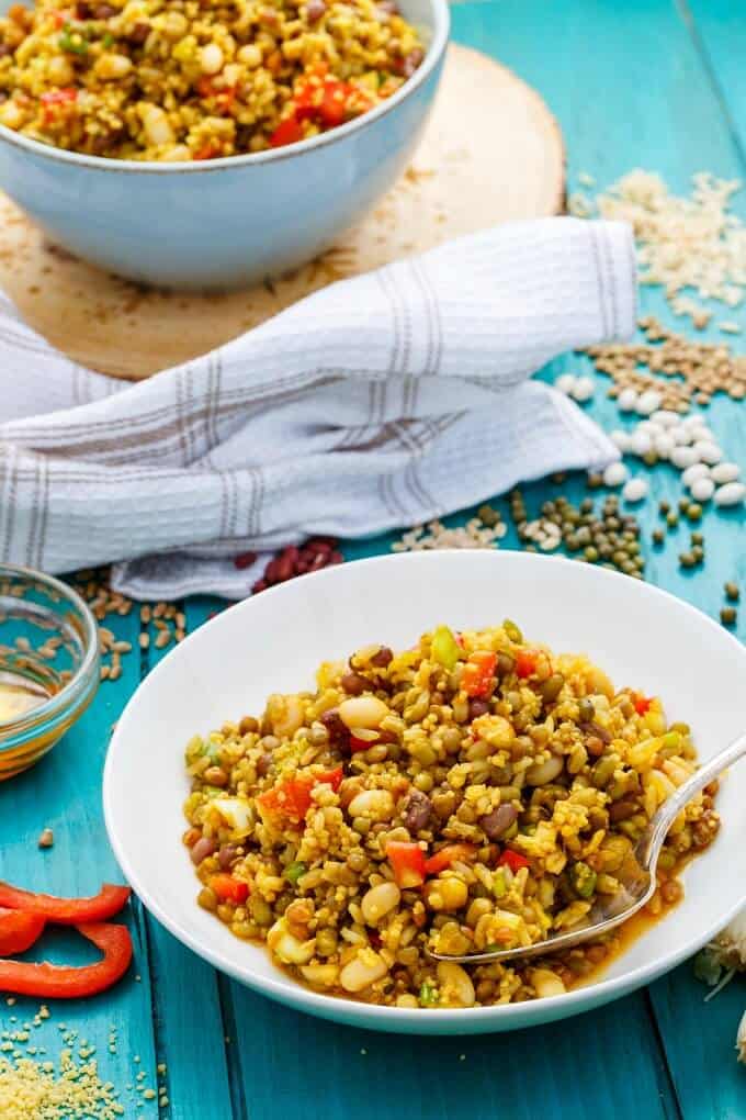 Summer Fresh Seven Grain Salad on white plate with spoon and in white bowl on wooden pad on blue table with many ingredients