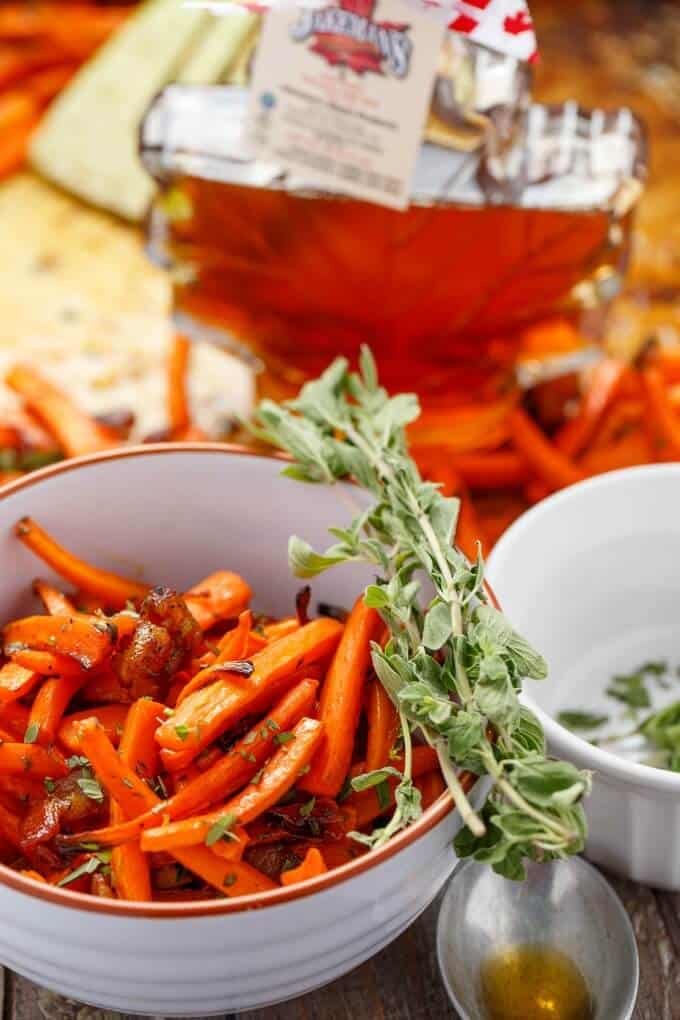 Maple Bacon Carrots in white bowl with herb, bottle of maple syrup in the background