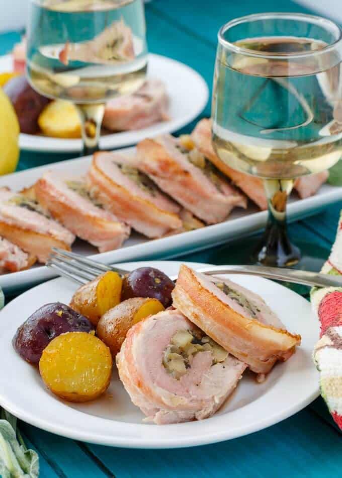 Bacon-Wrapped Apple Stuffed Pork Tenderloin on white plate with fork and potatoes. Tray with same dish, wine glass and cloth wipe on blue table