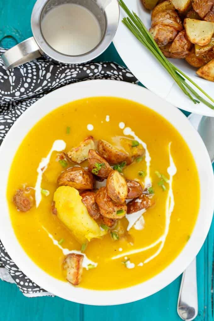 Roasted Apple Pumpkin Soup with Potatoes on white plate on blue table with spoon, plate with potatoes, metal jar and cloth wipe