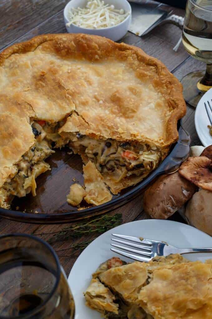 Roasted Mushroom Pot Pie on glass tray and white plate on table with  glasses of wine, fork, herbs, mushroom in bowl and on the table