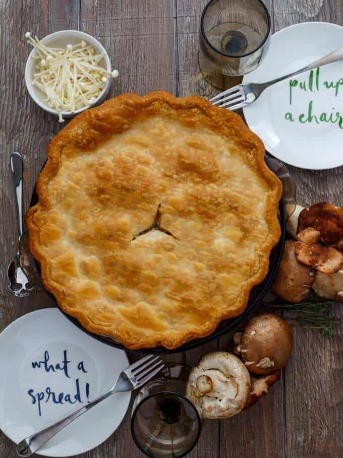 Roasted Mushroom Pot Pie on tray on wooden table with white plates, forsk, spoon, wine glasses, mushrooms in bowl and spilled on the table