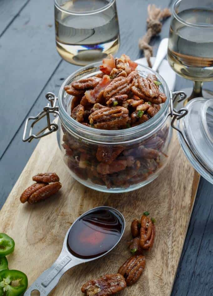 Bacon Jalapeno Pecans on wooden pad with pecans, jalapeno slices, spoon, wine glasses