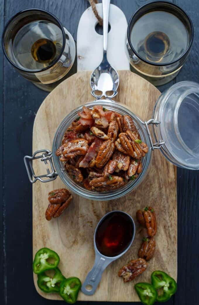 Bacon Jalapeno Pecans in glass jar on wooden pad with pecans, jalapeno slices, spoons, wine glasses