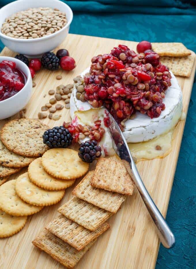 Cranberry Lentil Brie Bake on wooden pad with knife, crackers, cranberries, blackberries, bowls of lentils and cranberry sauce