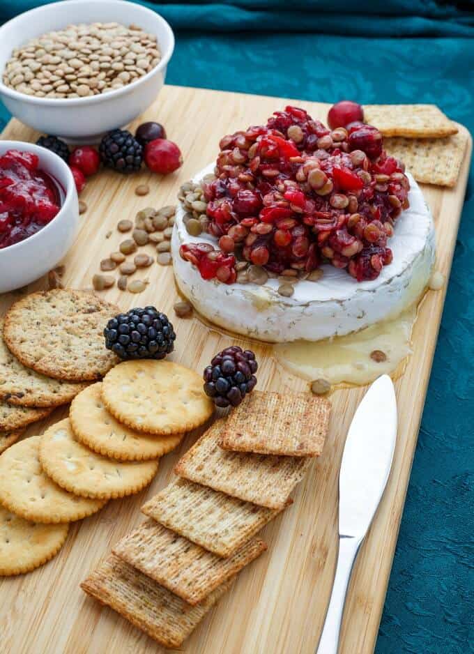 Cranberry Lentil Brie Bake on wooden pad with knife, crackers, raspberries, cranberries, bowl of fruits and lentils