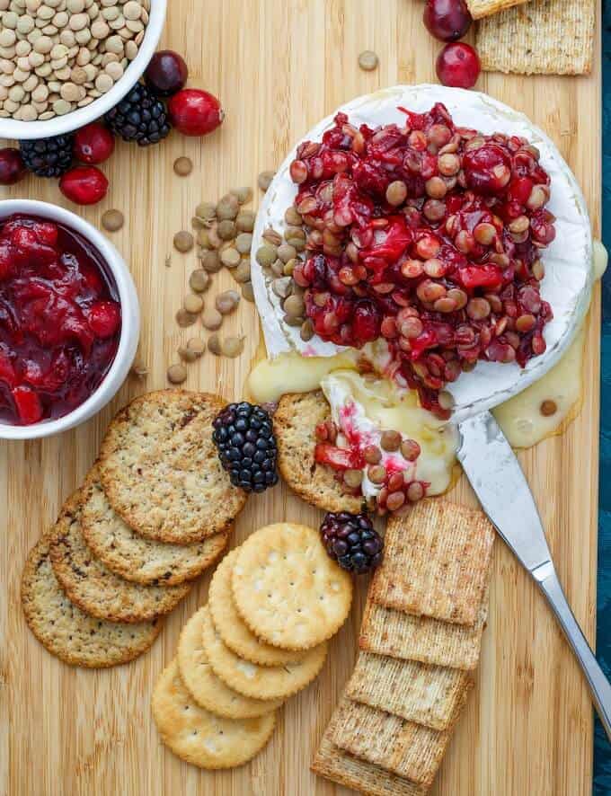 Cranberry Lentil Brie Bake on wooden pad with knife, crackers, bowl of lentils and bowls of sauce, blackberries, cranberries and lentils spilled around