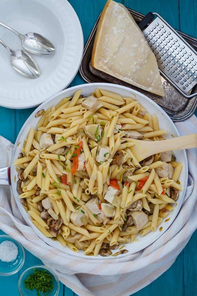 One Pan Creamy Chicken Pasta  in white bowl with wooden spatula. White plates with spoon, tray with cheese and grater and bowls with ingredients on blue table