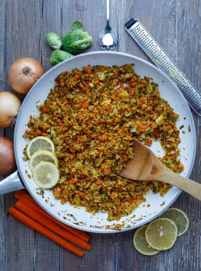 Shredded Lemon Brussels Sprouts Saute  on pan with wooden spatula on table with lemons slices, carrots, onions, cale, spoon, grater