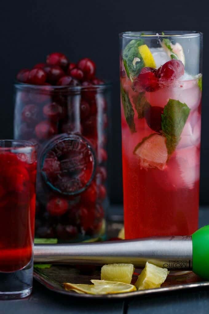 Christmas Cranberry Mojitos in glass cup on tray with lime, glass shot and glass jar full of cranberries in the background