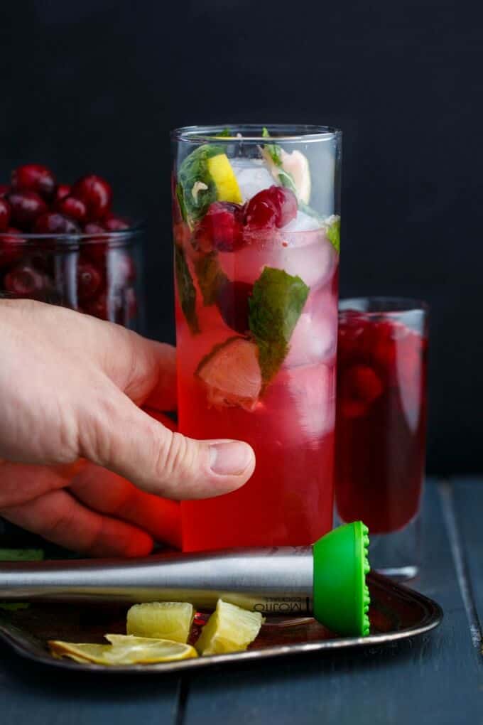 Christmas Cranberry Mojitos held by hand on gray table with lemon slices on tray, glass shot and jar of cranberries in the background