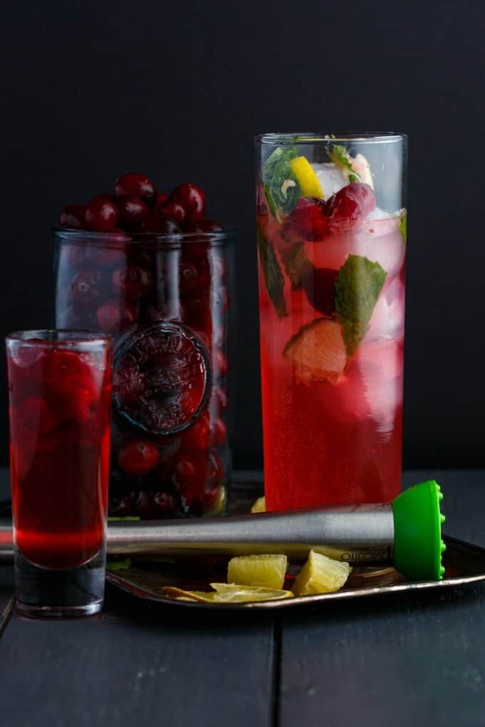 Christmas Cranberry Mojitos in glass cup and shot with limes on tray, glass jar full of cranberries