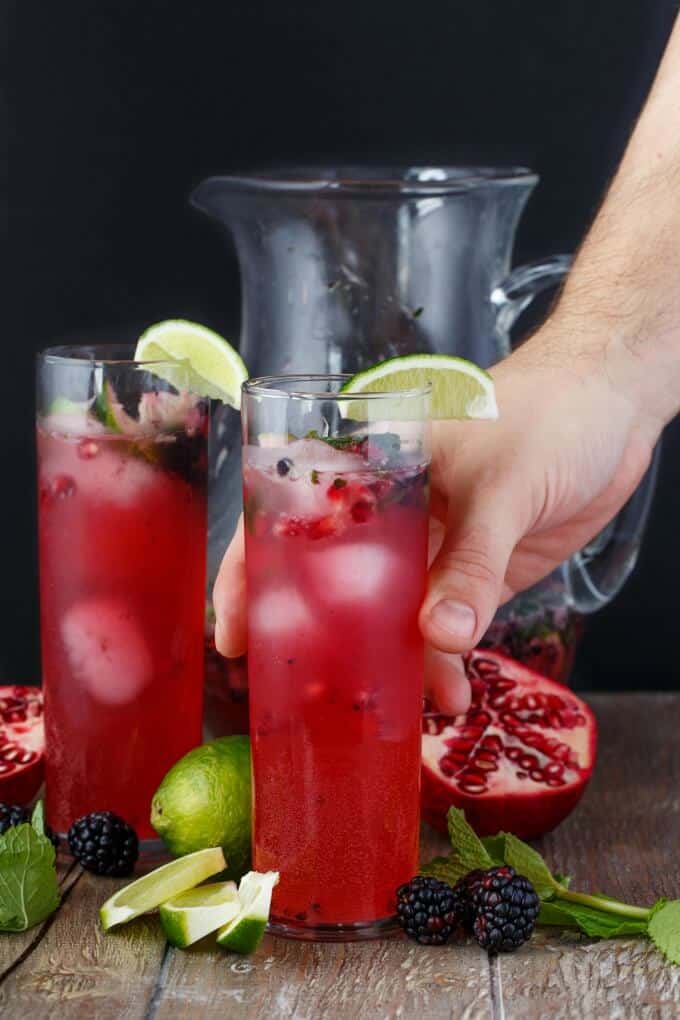 Pomegranate Blackberry Mojitos in glass pitcher and cups held by hand with lime slices, raspberries , herb, limes on the table