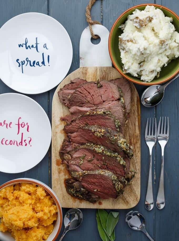 Slow Cooker Herb Butter Inside Round Roast on wooden pad on gray table with spoons, forks, bowls of side dishes, white plates