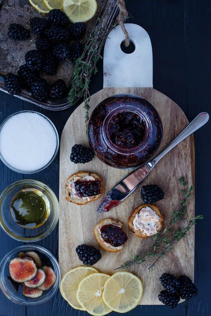 Blackberry fig jam on wooden pad with knife, snacks, lemons slices, blackberires, herd and bowls full of ingredients
