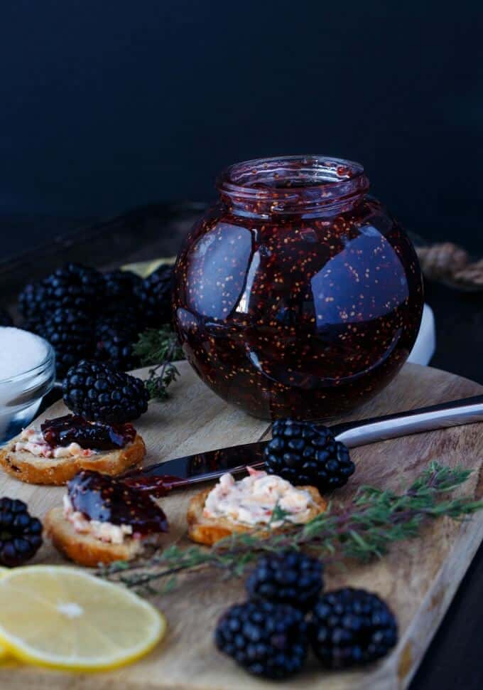 Blackberry fig jam in glasss jar on wooden pad with knife, blackberries, lemons slices, herb, snacks