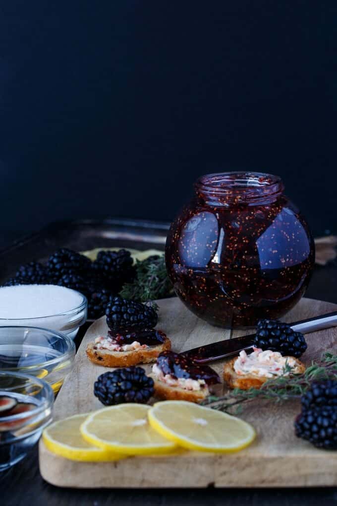 Blackberry-Fig Jam in glass jar on wooden pad with blackberries, lemons, knife, snack and bowls full of ingredients