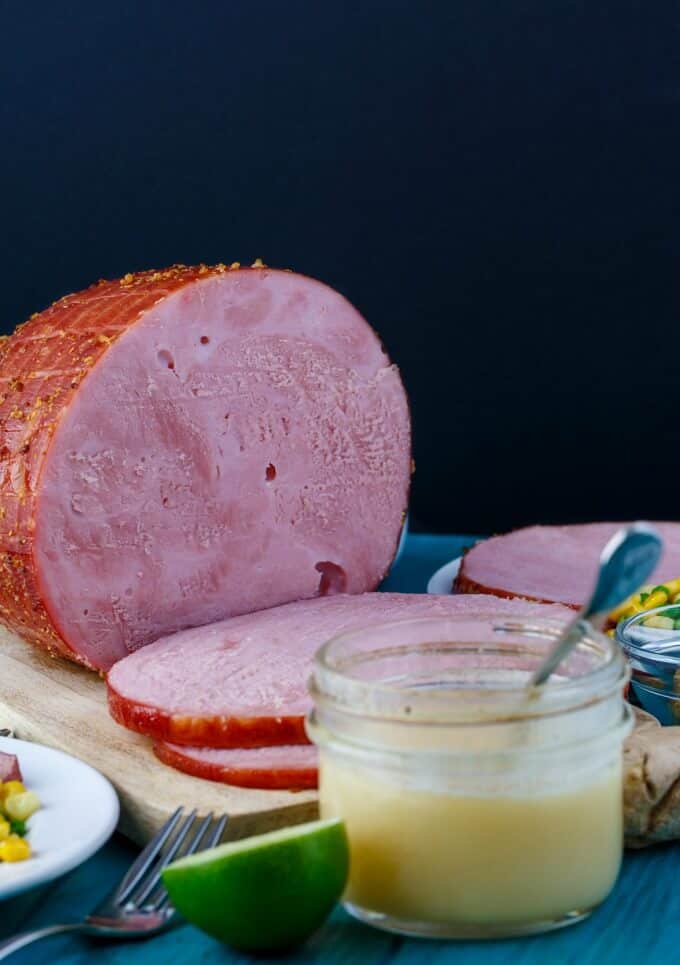 Slow Cooker Honey-Garlic Ham  sliced on wooden pad on blue table with lime, fork jar of sauce with spoon