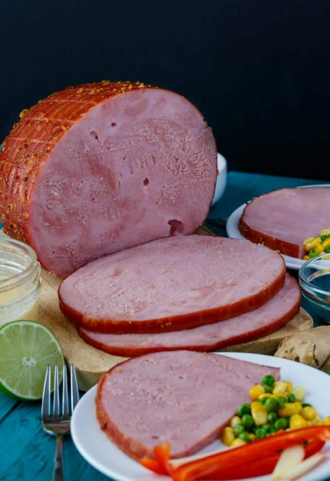 Slow Cooker Honey-Garlic Ham sliced on wooden pad and on white plate with vegetables on blue table with fork and lime