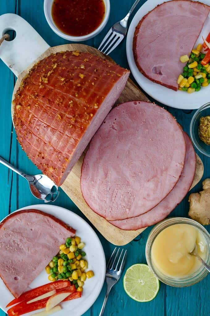 Slow Cooker Honey-Garlic Ham slcied on wodoen pad and on white plates with vegetables on blue table with spoon, forks, lime and jar of sauce with spoon