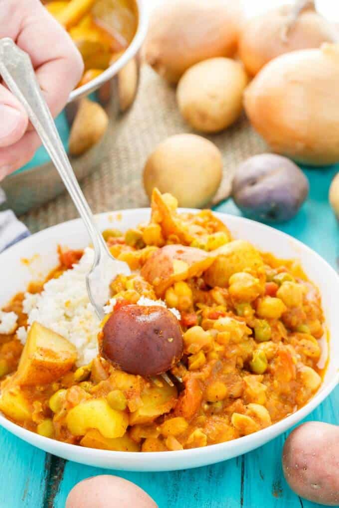 Vegan Potato Curry over Cauliflower Rice on white plate with fork held by hand, on blue table with spilled potatoes