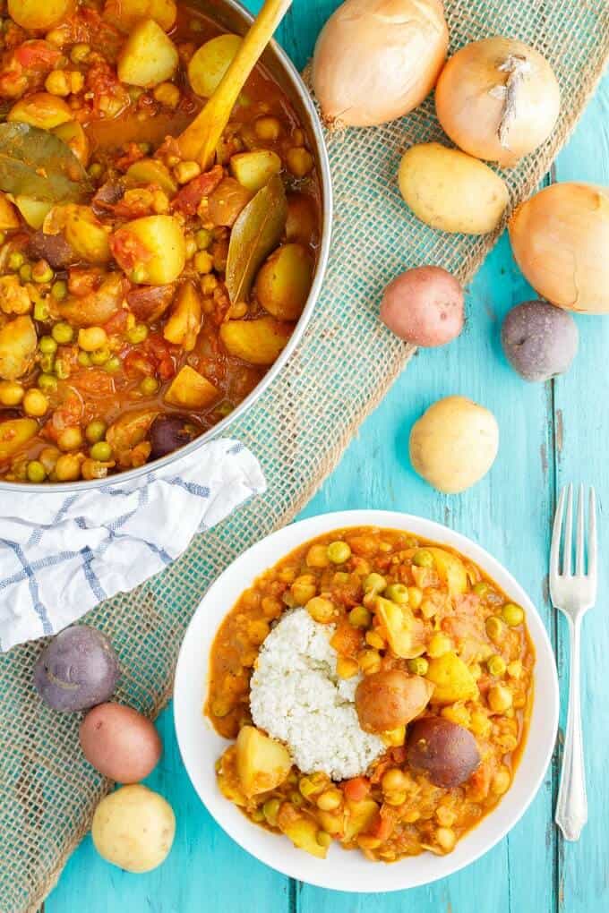 Vegan Potato Curry over Cauliflower Rice on white plare on blue table with fork, potatoes , onions, pan with same dish and wooden spatula