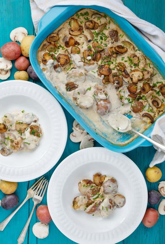 Cream of Mushroom Soup Potatoes in blue casserole with spoon, on white plates. Scattered potatoes, mushrooms on blue table with forks
