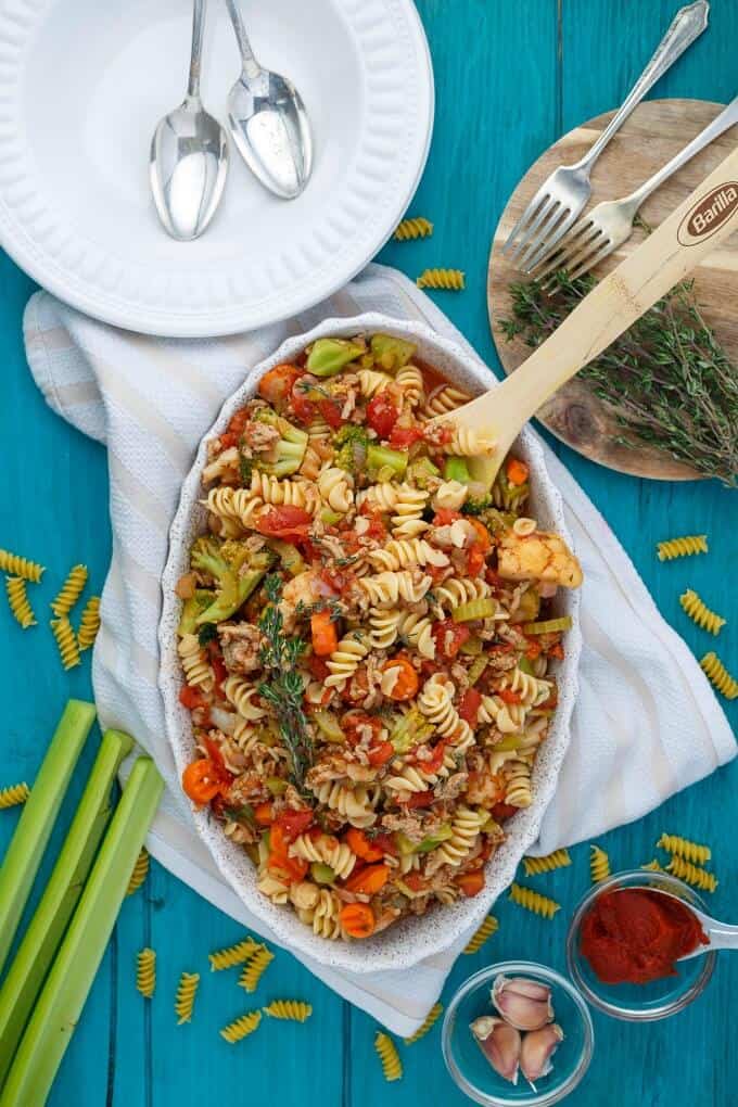 Loaded Vegetable Pasta in Casserole with wooden spatula on table with white plate with spoon, wooden pad with herbs and forks, cloth wipe and pasta, vegetables spilled on blue table with garlic and sauce