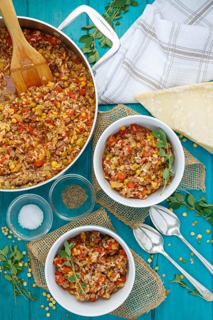 Split Pea Lentil Casserole in pad with wooden spatula and in white bowls. Spoon, ingredients in bowls, cloth wipe, herbs, scattered lentils and cheese on blue table