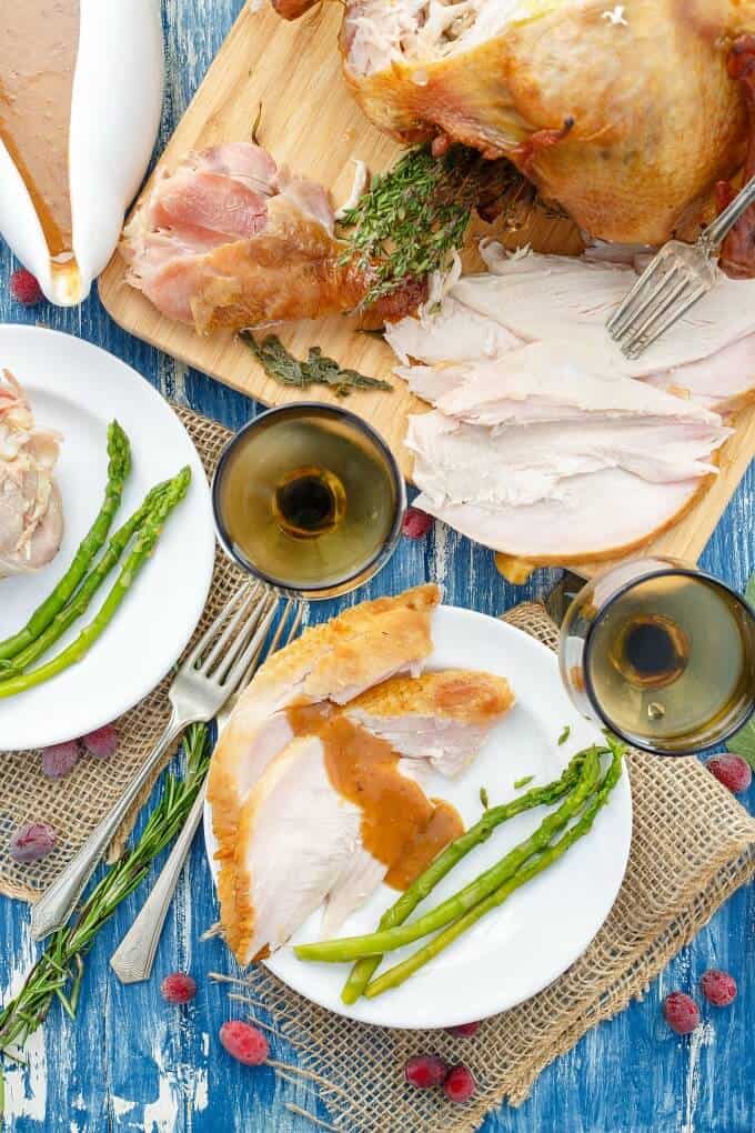 Smoked Turkey on woodenn pad and white plates with vegetables on blue table with wine glasses and bowl of sauce, forks