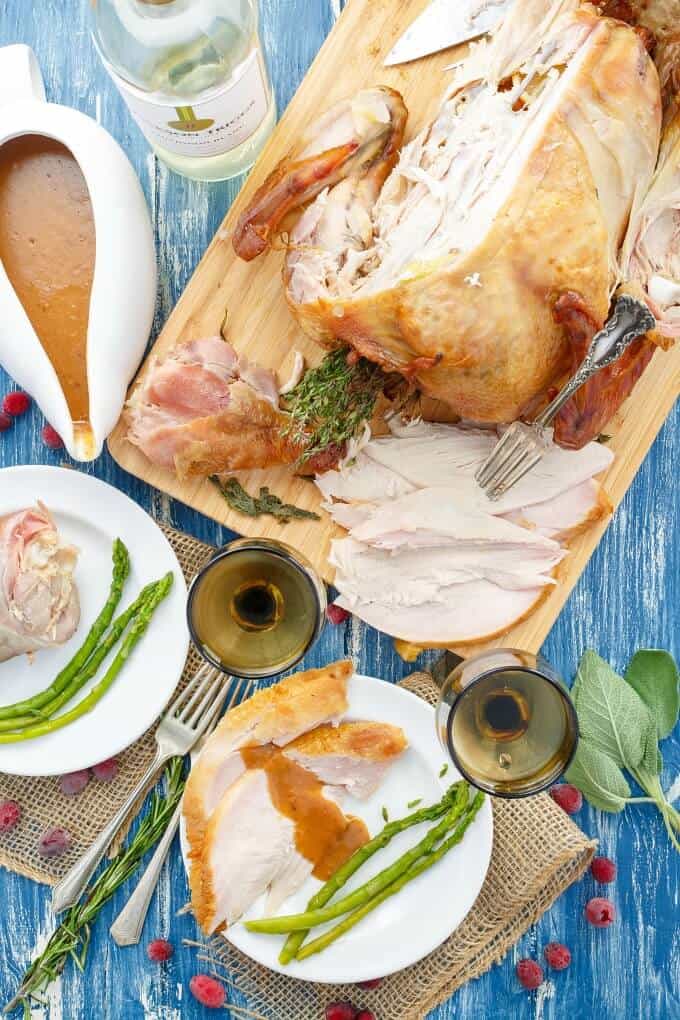 Smoked Turkey on wooden pad and white plates with vegetables on blue table with forks, bottle of wine, wine glasses, bowl of sauce