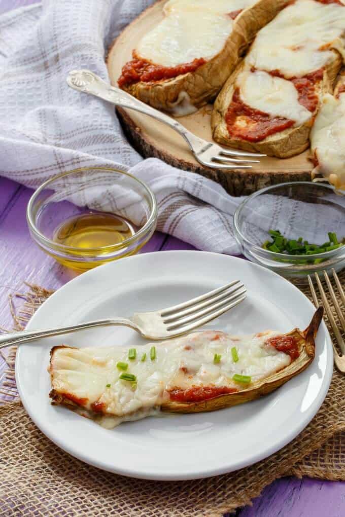 Baked Eggplant Pizza Crust Slices on white plate and wooden pad wit forks, ingredients in small bowls on purple table