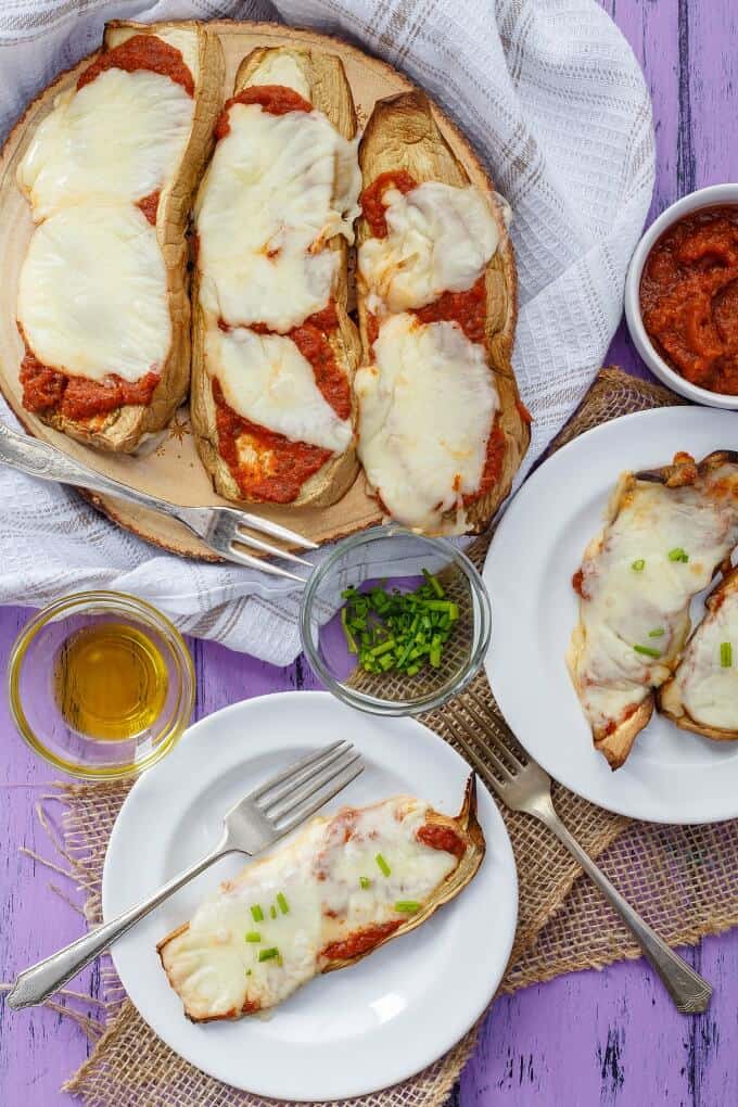 Baked Eggplant Pizza Crust Slices on white plates and on wooden pad with forks, ingredients in small bowls on purple table