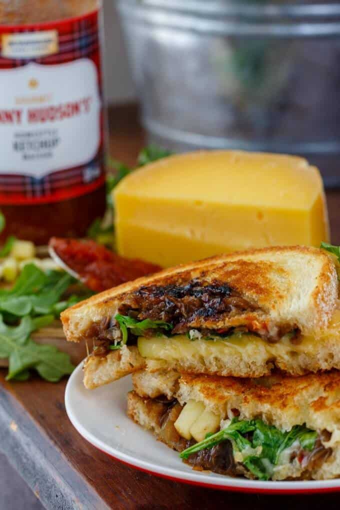 Caramelized Onion-Mushroom Grilled Cheese Sandwich with Apple on white plate, cheese, herbs and sauce in the background