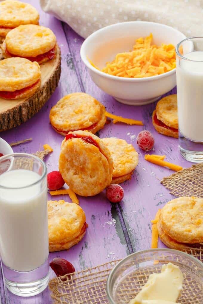 Cheesy Leftover Cranberry Sauce Cookies on wooden pad and puple table with glasses of milk, bolw of cheese, butter and scattered cranberries