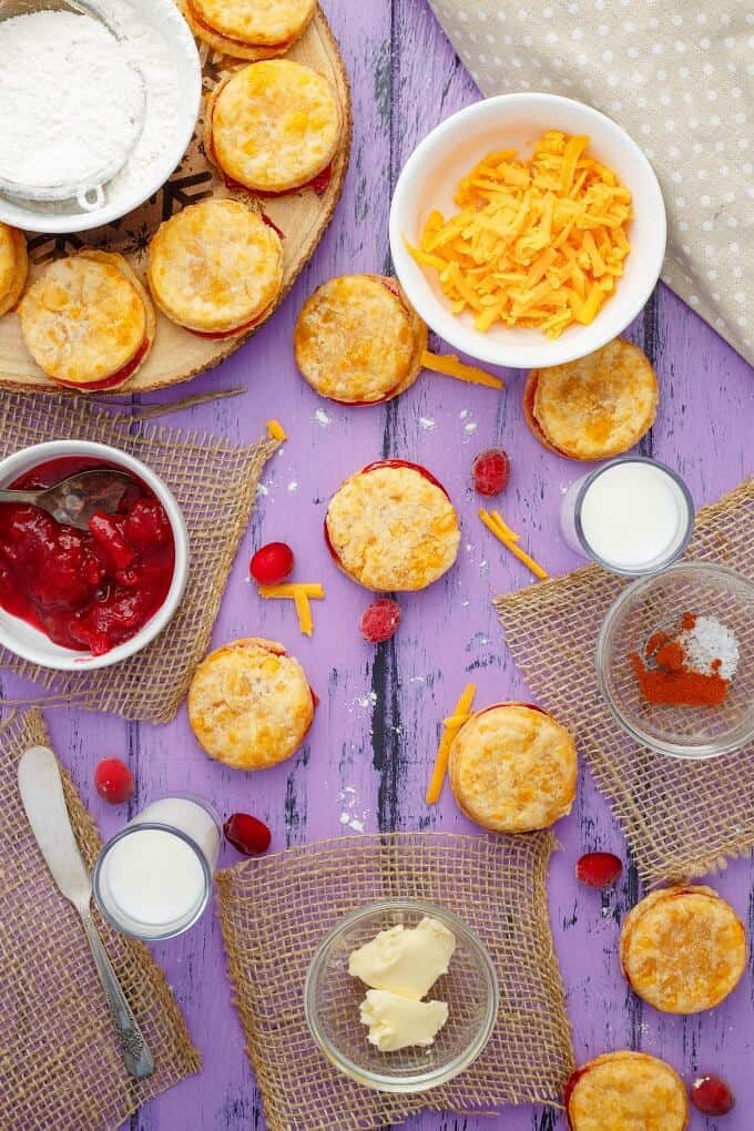 Cheesy Leftover Cranberry Sauce Cookies on wooden pad and purple table with bowl of cheese, bowl of cranberry sauce, knife, bowls of ingredients and spilled cranberries