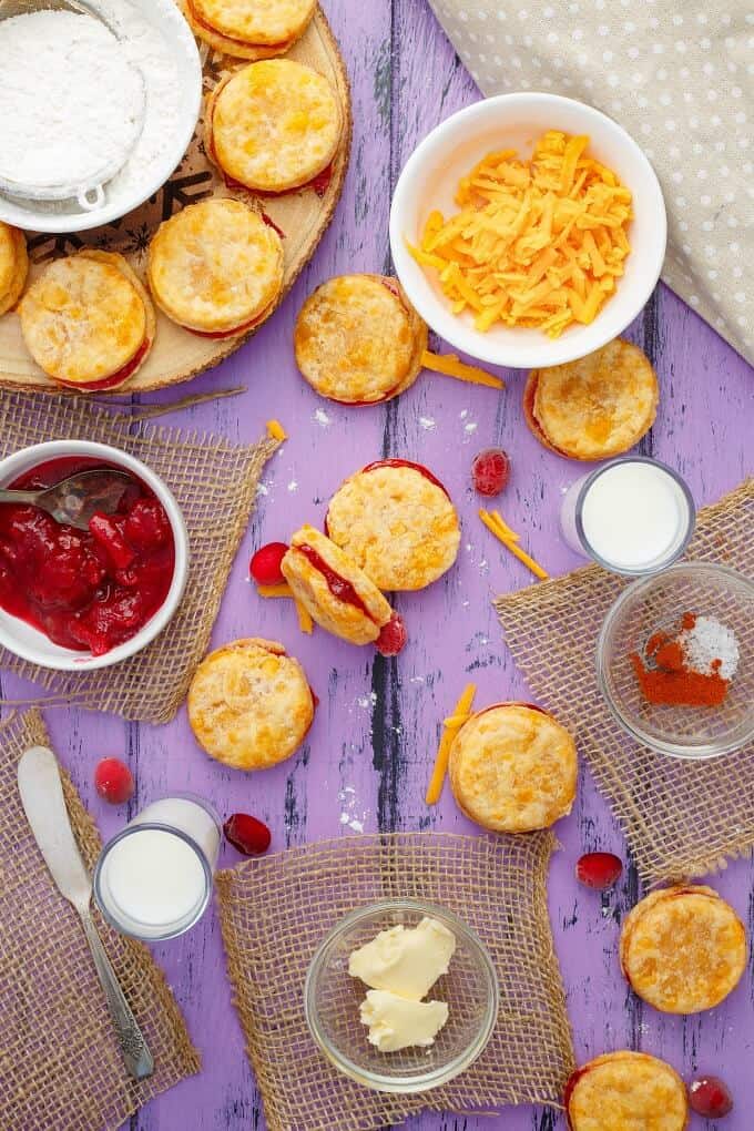 Cheesy Leftover Cranberry Sauce Cookies on wooden pad and purple table with bowls of cheese, sauce, ingredients, knife, scattered cranberries around and glasses of milk