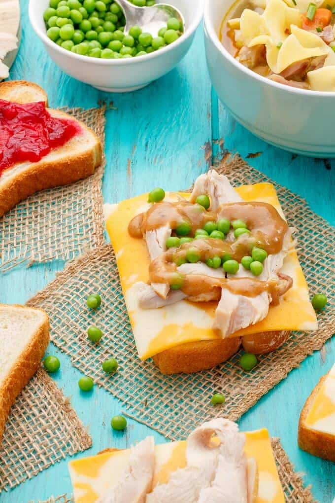 Open-Faced Turkey Sandwiches (Using Leftover Turkey) with peas on blue table next to sandwich with jam, bowl full of dish, bowl of peas and scattered peas around