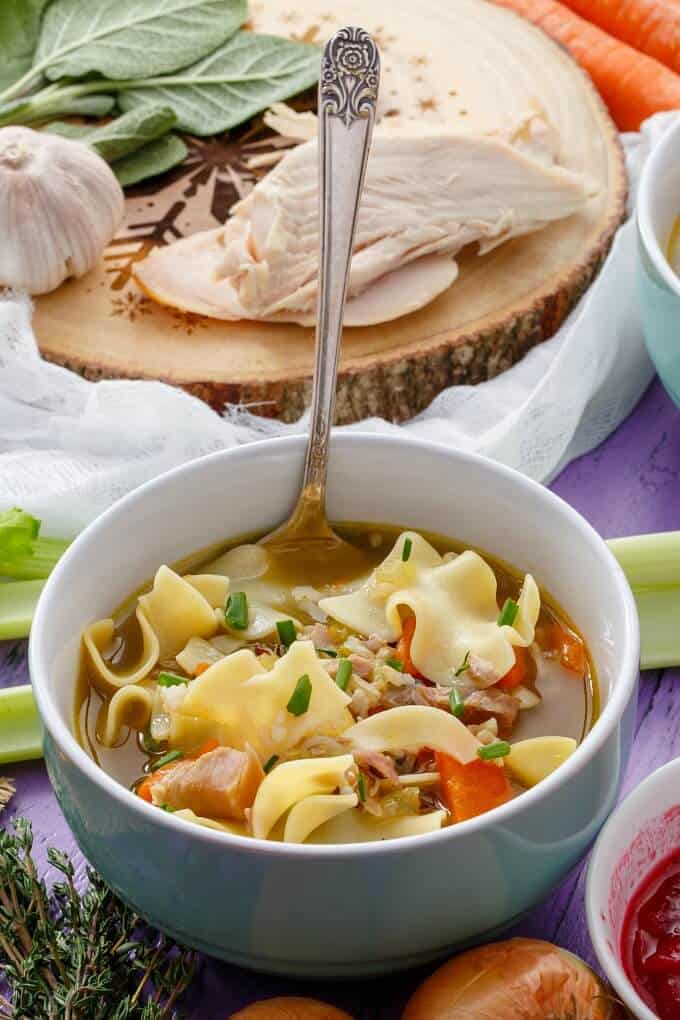 Turkey Noodle Soup  in blue bowl with spoon, turkey,garlic, herbs on wooden pad, veggies on the table