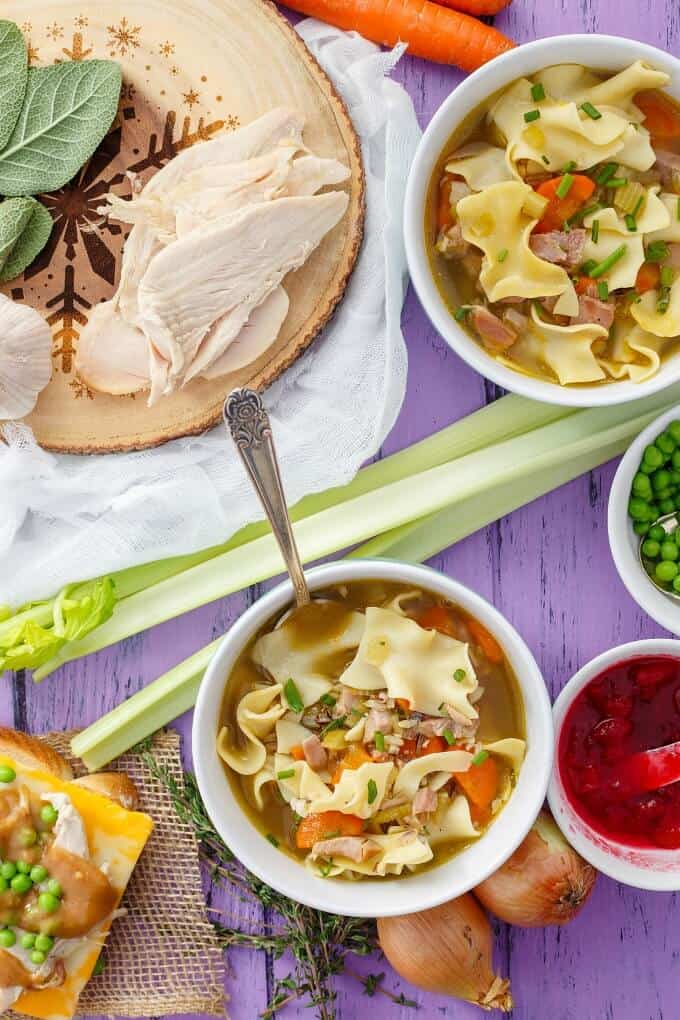 Turkey Noodle Soup in blue table with spoon on purple table with asparagus, onions, pears in bowl, sauce in bowl, wooden pad with leftover turkey