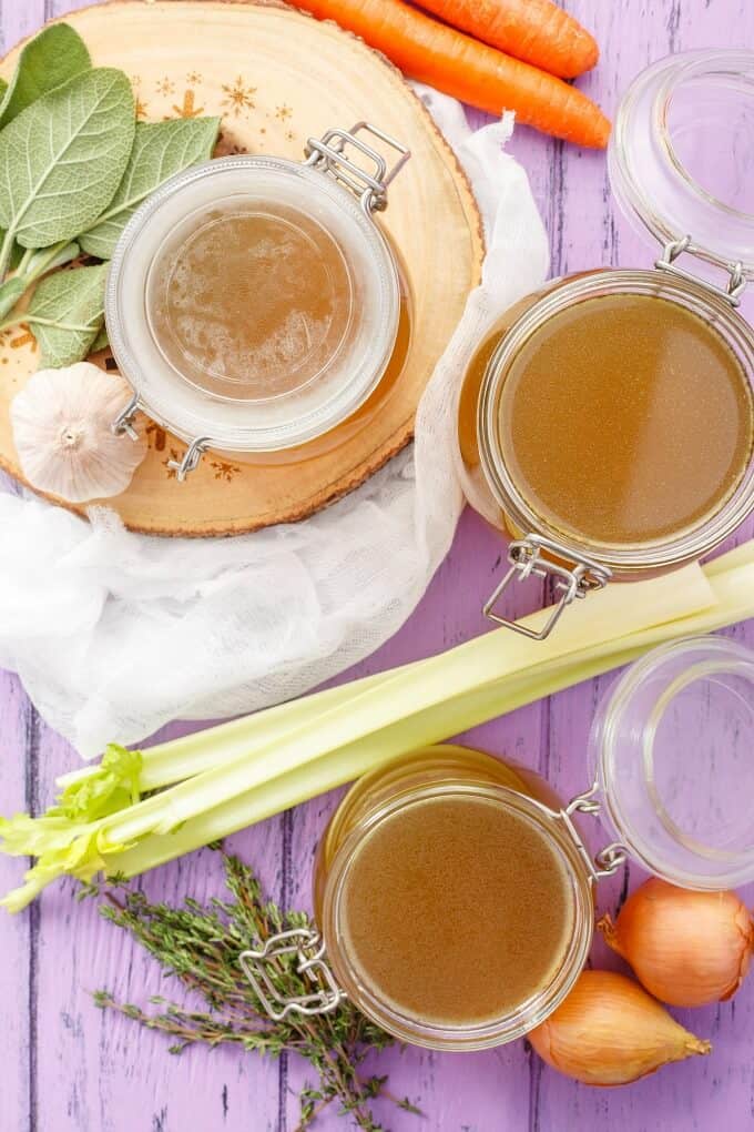 Homemade Turkey Stock in glass jars on wooden pad and purple table with carrots, onions, garlic and herbs