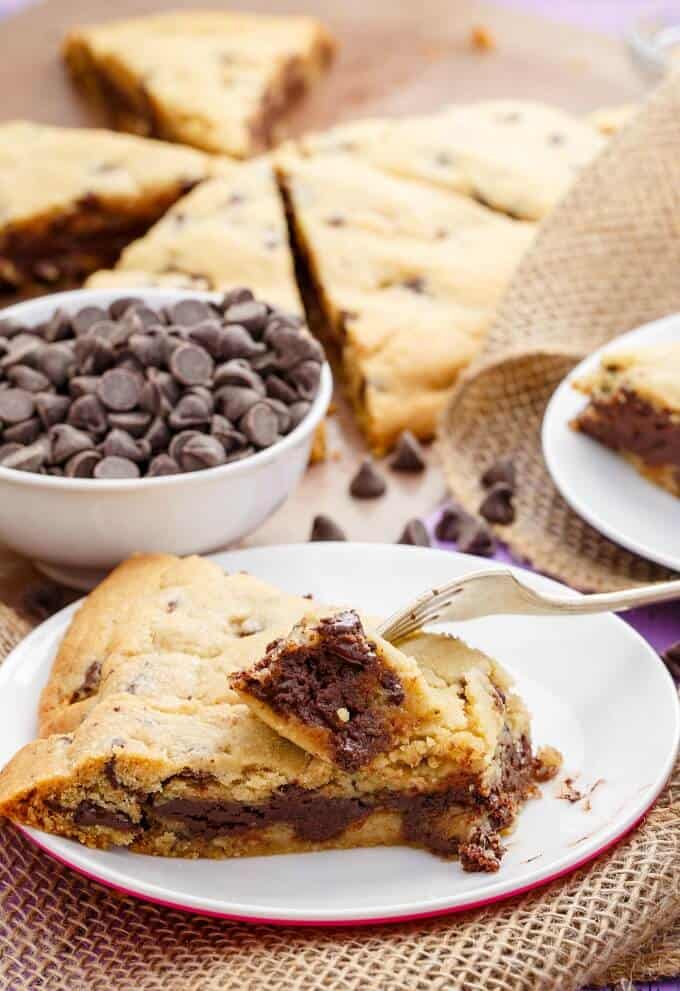 Giant Fudge Stuffed Cookie on white plate with fork next to white bowl of chocolate chips and slices of cake in the background