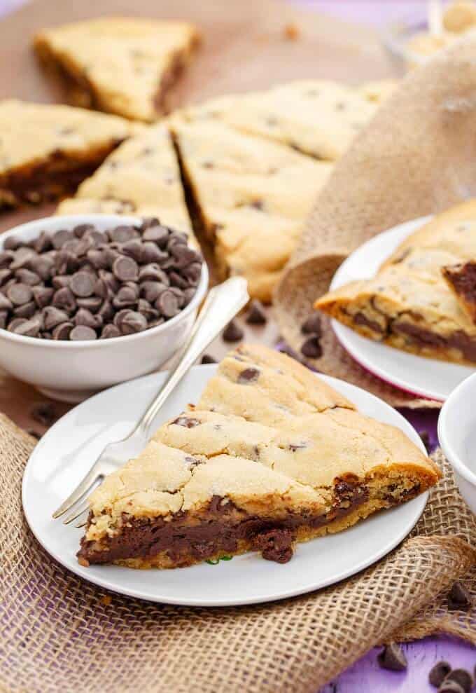 Giant Fudge Stuffed Cookie on white plates with fork, on baking paper, bowl full of chocolate chips, chips scattered on purple table