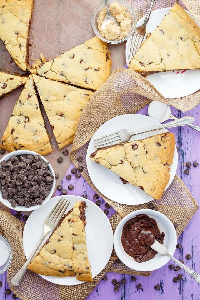 Giant Fudge Stuffed Cookie on white plates with forks and on purple table with chocolate chips in white bowl and chocolate pudding in white bowl with knife, chocolate chips scattered on purple table
