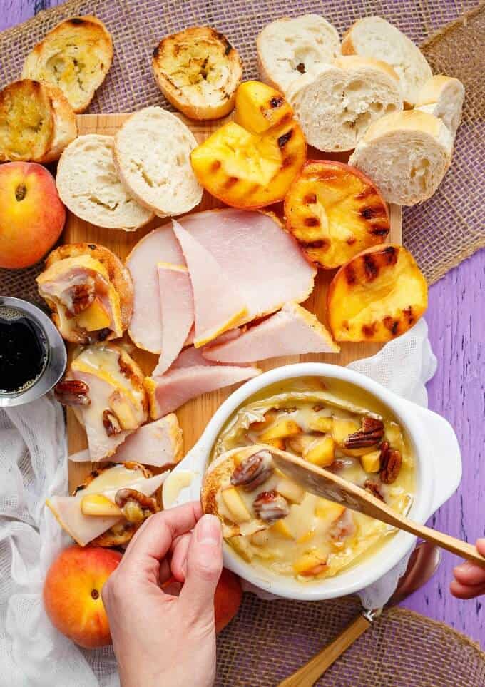 Grilled Maple-Butter Peach Camembert with Maple Pecans & Peameal Bacon in white bowl mixed by wooden sptual held by hand, all the ingredients on the table