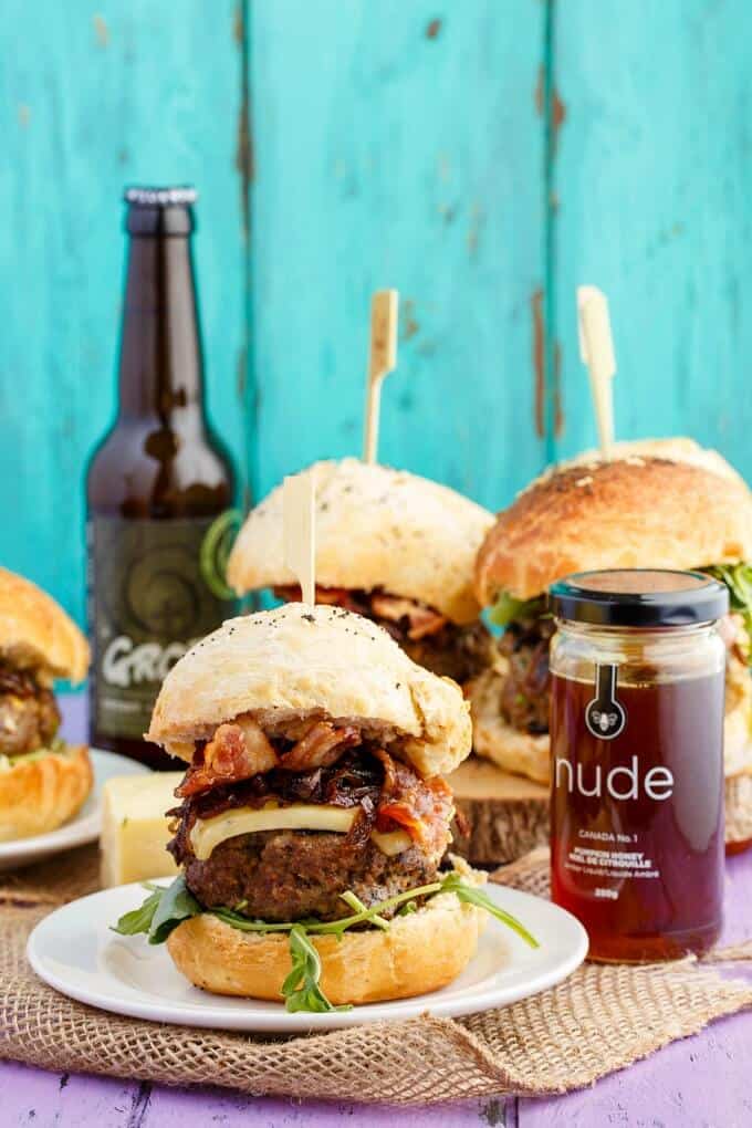 Homemade Honey-Garlic Hamburgers with Maple-Bacon Caramelized Onions on white plate and wooden pad. Honey jar and beer bottle on purple table